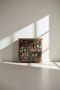 A small wooden cabinet or chest covered in raised alphabet blocks, photographed in a minimalist room with sunlight casting strong shadows on a white wall and floor.