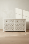 An off-white wooden six-drawer dresser with intricate carved patterns on the drawer fronts, shown in a minimalist room with light wood floors and a textured wall.