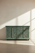 An ornate carved wooden sideboard cabinet in a distressed teal-green finish, set against a minimalist beige wall with soft window shadow patterns.