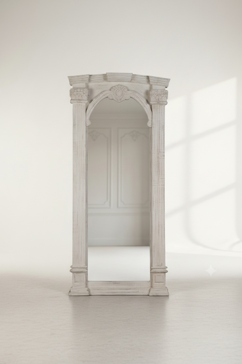 A tall, narrow white arched mirror with decorative classical pillars and a small crest at the top, photographed in a bright minimalist room with a pale floor and soft window shadows.