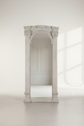 A tall, narrow white arched mirror with decorative classical pillars and a small crest at the top, photographed in a bright minimalist room with a pale floor and soft window shadows.