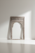 An ornate, rectangular Moroccan-style carved wooden arch frame in a weathered light gray finish, leaning against a minimalist white wall with soft sunlight casting a shadow.
