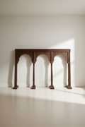 An ornate dark brown wooden console table with three arched supports and carved details, set against a minimalist off-white wall and floor with soft sunlight casting shadows.